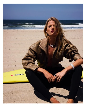 Woman in brown leather jacket and black pants seated on sand at a beach with a surfboard in the background