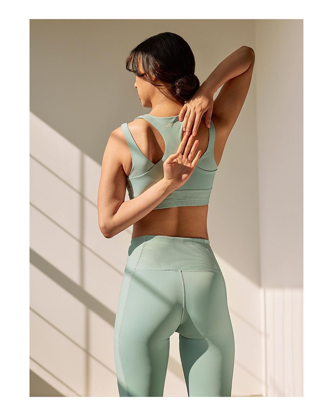 Woman in sportswear stretches her arm behind her back. She is indoors in front of a light beige wall with sunlight.