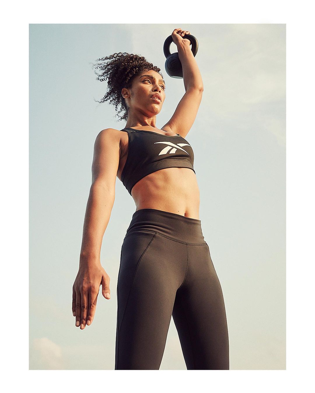 Athletic woman in sportswear holding kettlebell overhead against a clear sky. Fitness workout, outdoor training, healthy lifestyle.