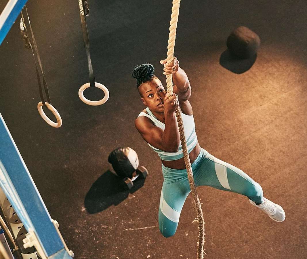 Athletic woman climbs thick rope in gym with rings, weights, and rubber floor, wearing teal and white sports top and leggings.