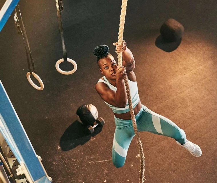 Athletic woman climbs thick rope in gym with rings, weights, and rubber floor, wearing teal and white sports top and leggings.