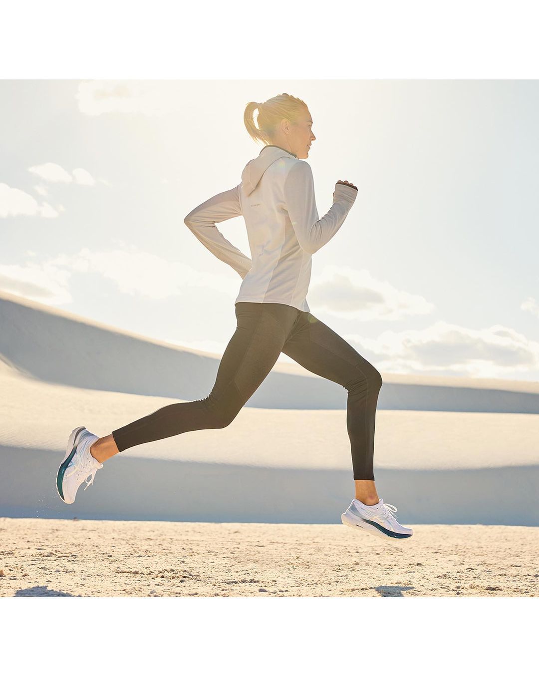 Woman running in athletic wear on tan, sandy dunes under bright sunlight, wearing black leggings, white running shoes and light grey jacket.