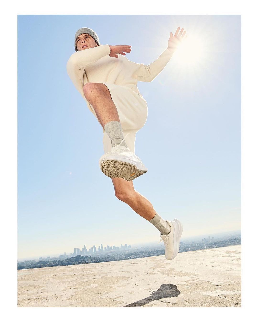 Athletic man mid-leap in cream-colored apparel and running shoes, against a backdrop of blue sky and distant cityscape.
