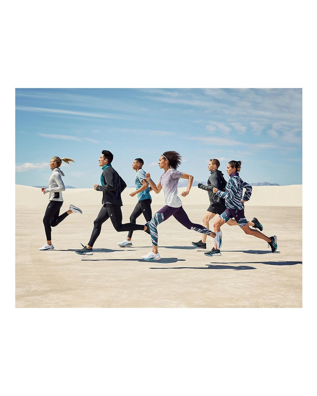 A group of seven people are running outdoors in athletic wear on a sunny day. They are on a flat sandy surface with a blue sky overhead.