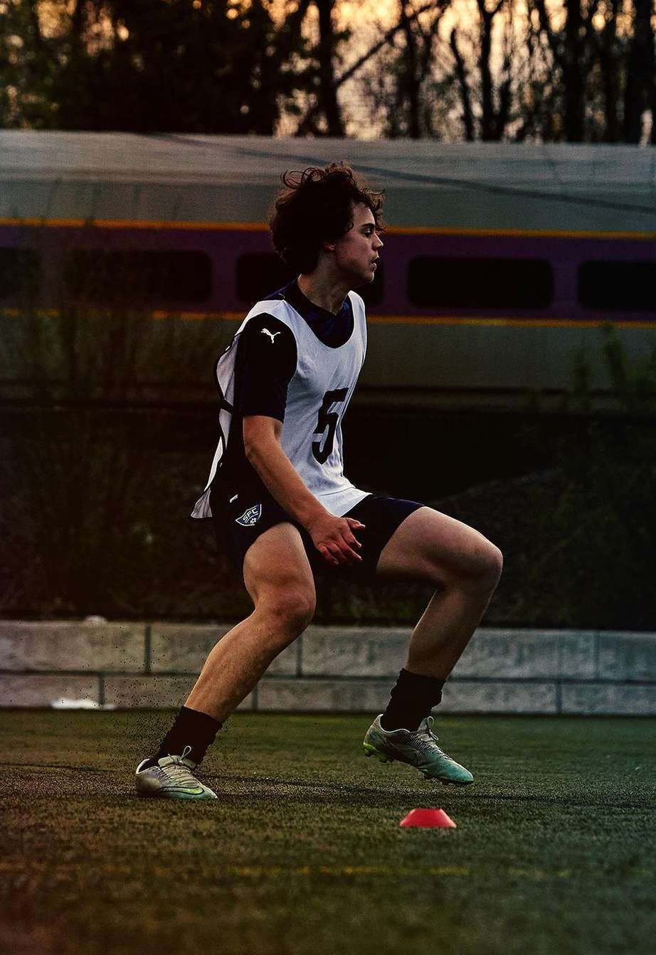 An athlete with curly dark hair in athletic gear is sprinting on an artificial-turf field with a red cone and train in the background.