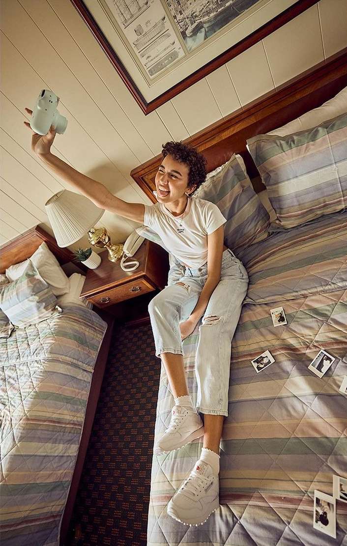 A young adult woman sits on a bed in a bedroom, holding a camera up to take a selfie, with other instant photos scattered on the bed.