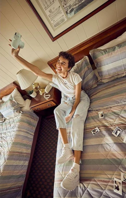 A young adult woman sits on a bed in a bedroom, holding a camera up to take a selfie, with other instant photos scattered on the bed.