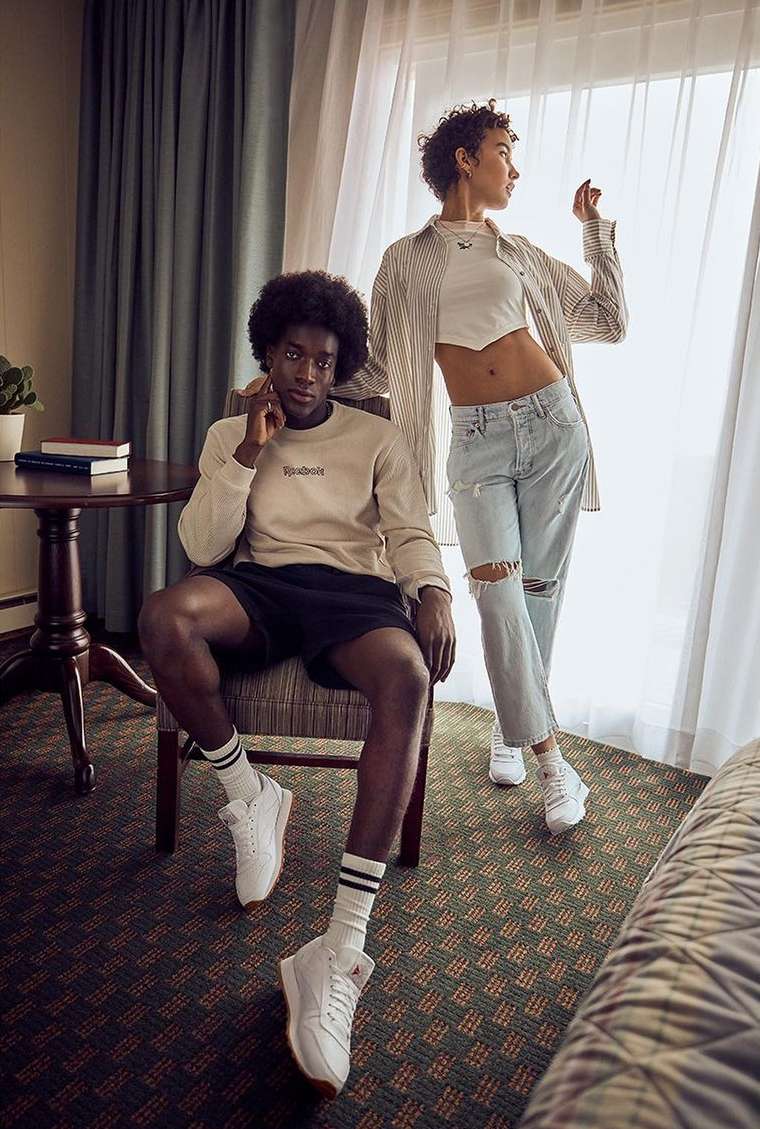 Two fashion models wearing casual attire, posed in a hotel room with natural window lighting, showcasing a modern, relaxed style indoors.