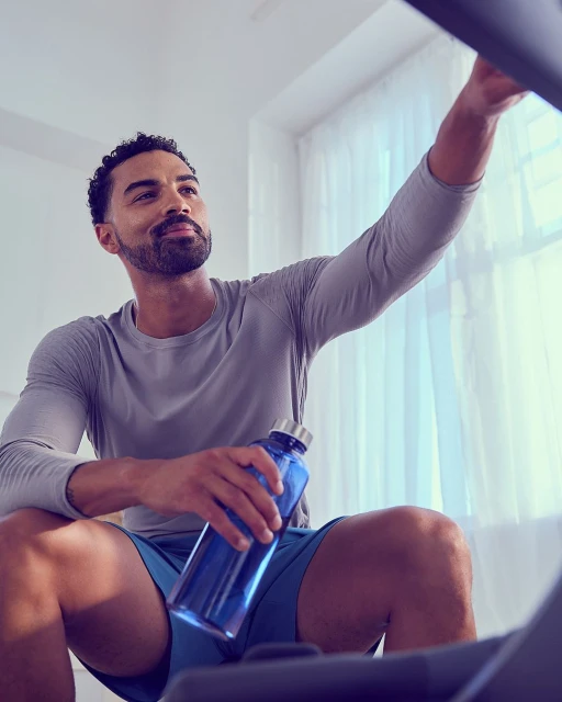 Athletic man in grey long-sleeve shirt sits by exercise equipment with a blue water bottle in hand and window curtains in the background.