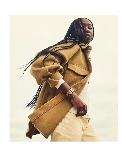 Dark-skinned woman in a tan coat and light trousers looking away from the camera in a relaxed pose against a soft light background.