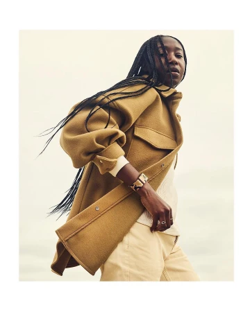 Dark-skinned woman in a tan coat and light trousers looking away from the camera in a relaxed pose against a soft light background.