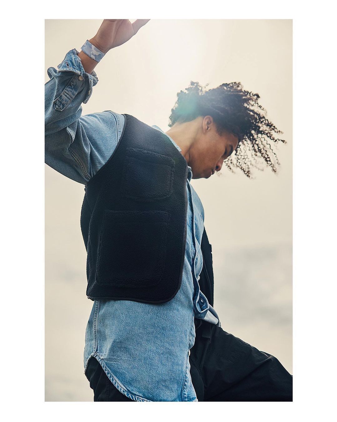 Man wearing a black fleece vest over a light denim shirt against a bright sky background in London by David Salafia