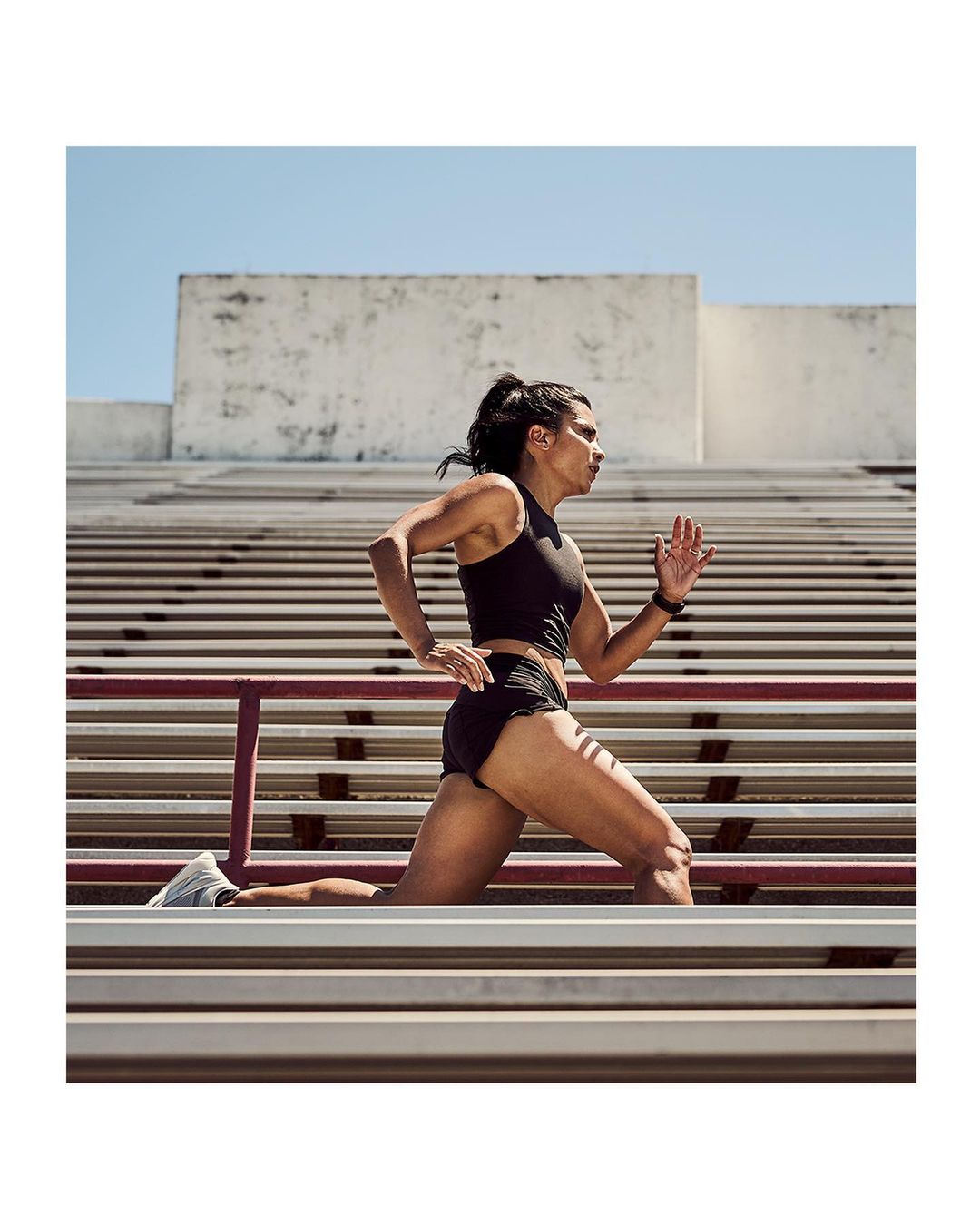Athletic woman runs on outdoor stadium steps in sports bra and shorts, viewed from a low angle on a sunny day.
