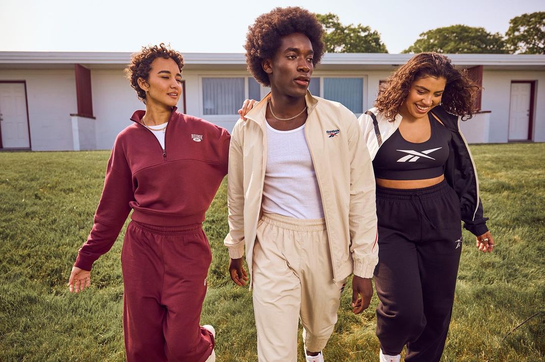 Three diverse adults in Reebok tracksuits walk on green grass in front of a light-colored building, showcasing fashion and lifestyle.