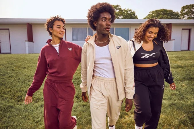 Three diverse adults in Reebok tracksuits walk on green grass in front of a light-colored building, showcasing fashion and lifestyle.