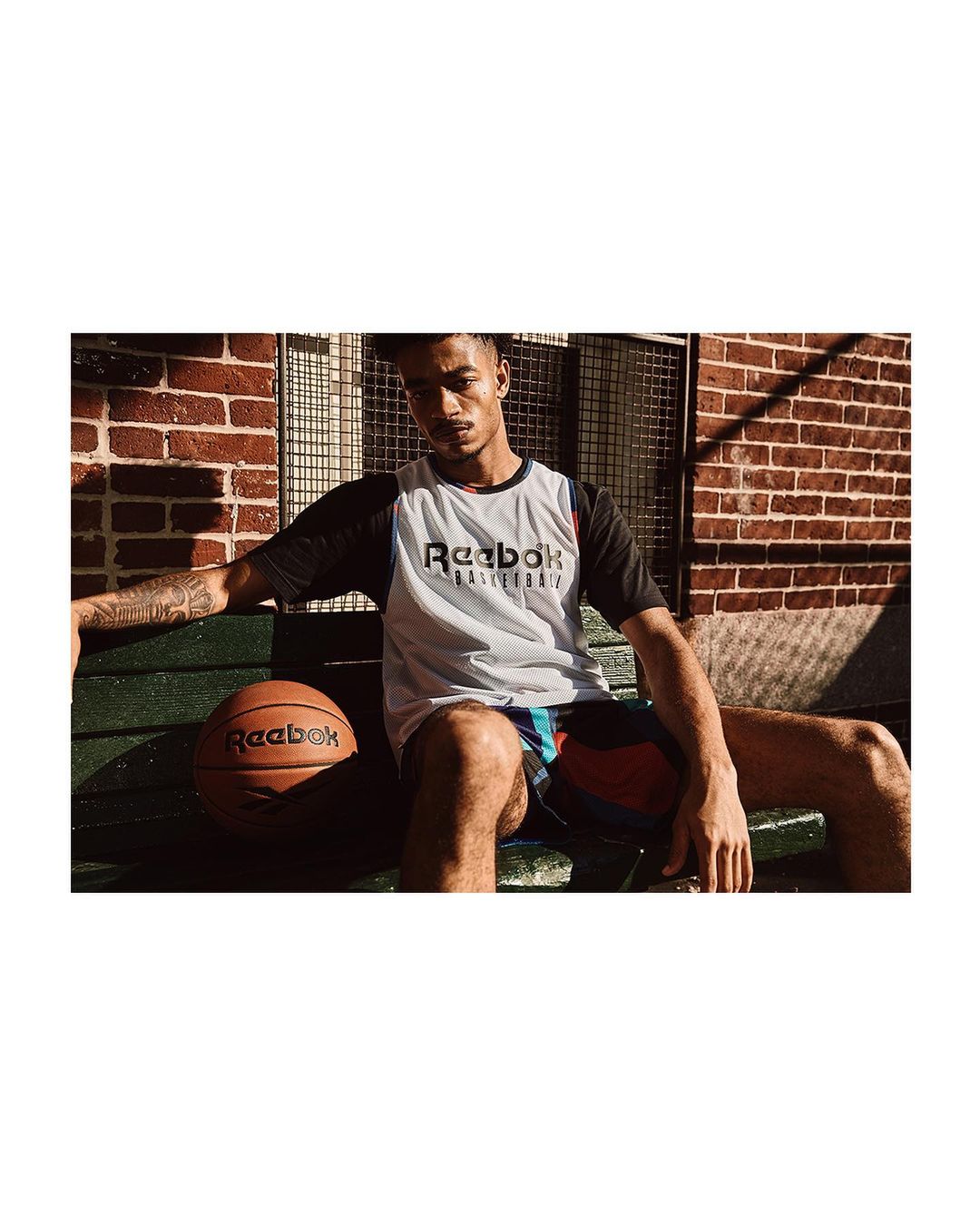 A young adult male sits on a green bench with a basketball next to him. He wears a Reebok jersey and shorts with a brick wall background.
