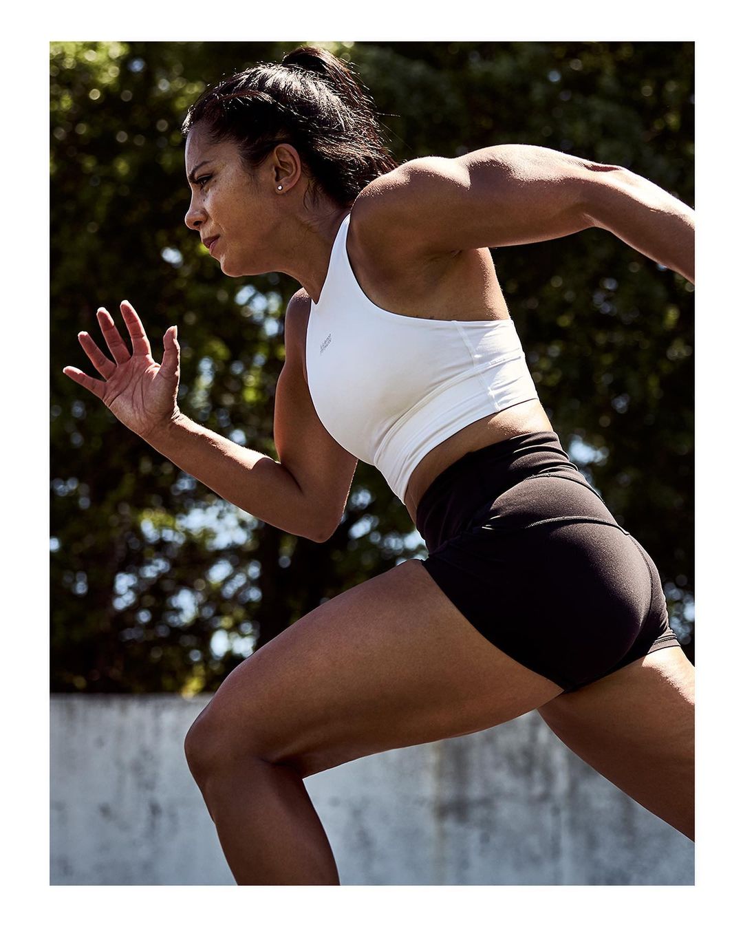 Athletic woman in white sports bra and black shorts running outdoors against a blurred green foliage background