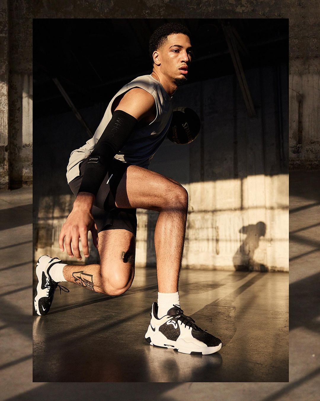 Male athlete in sportswear crouches while holding a basketball in a gritty indoor environment with strong directional lighting.