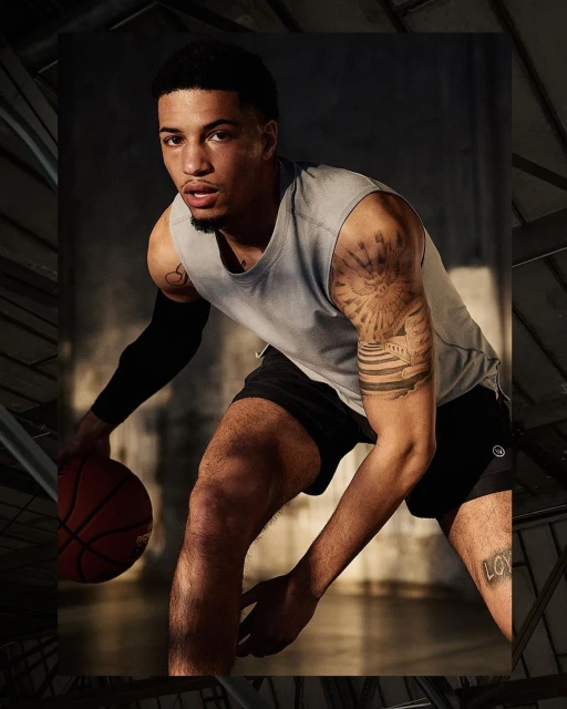Athletic Black male in tank top dribbling basketball indoors, sleeve tattoo art, dark background, fitness lifestyle, sports action.