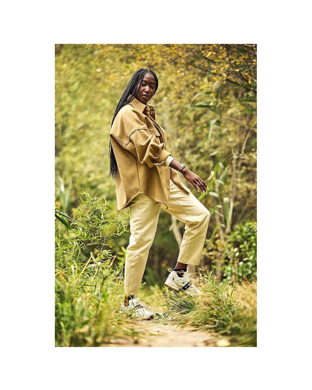 An adult woman with long dark braids walks on a dirt path in a park, wearing a brown overshirt, pale yellow pants and neutral-toned sneakers.