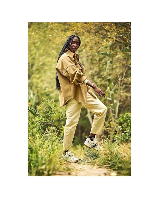 An adult woman with long dark braids walks on a dirt path in a park, wearing a brown overshirt, pale yellow pants and neutral-toned sneakers.
