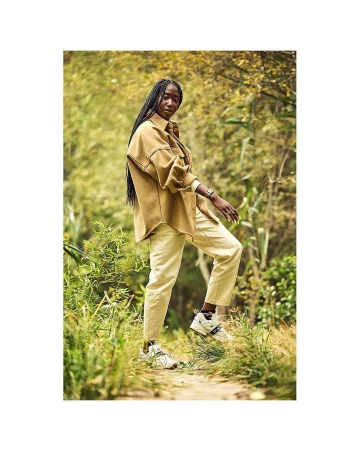 An adult woman with long dark braids walks on a dirt path in a park, wearing a brown overshirt, pale yellow pants and neutral-toned sneakers.