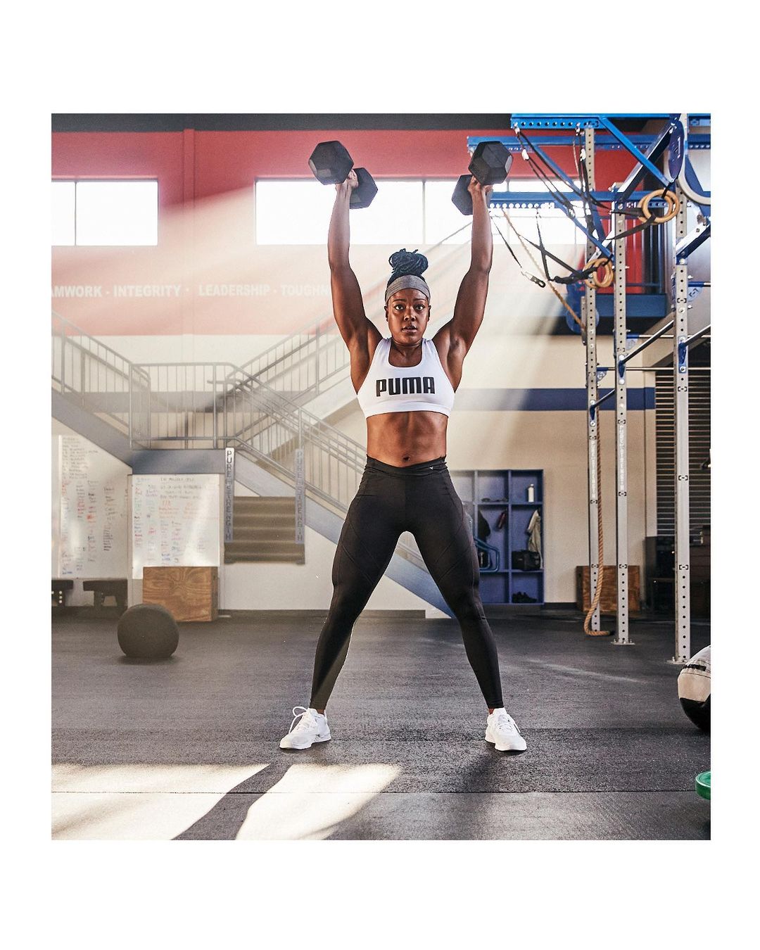 Athletic woman in sports bra and leggings lifting dumbbells overhead in a gym setting, showcasing strength and fitness.