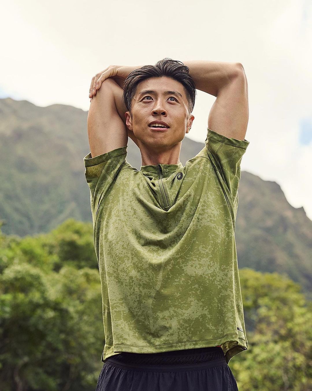 Athletic Asian man stretching arms in a green camo shirt outdoors with mountains and cloudy sky in the background, fitness and lifestyle.