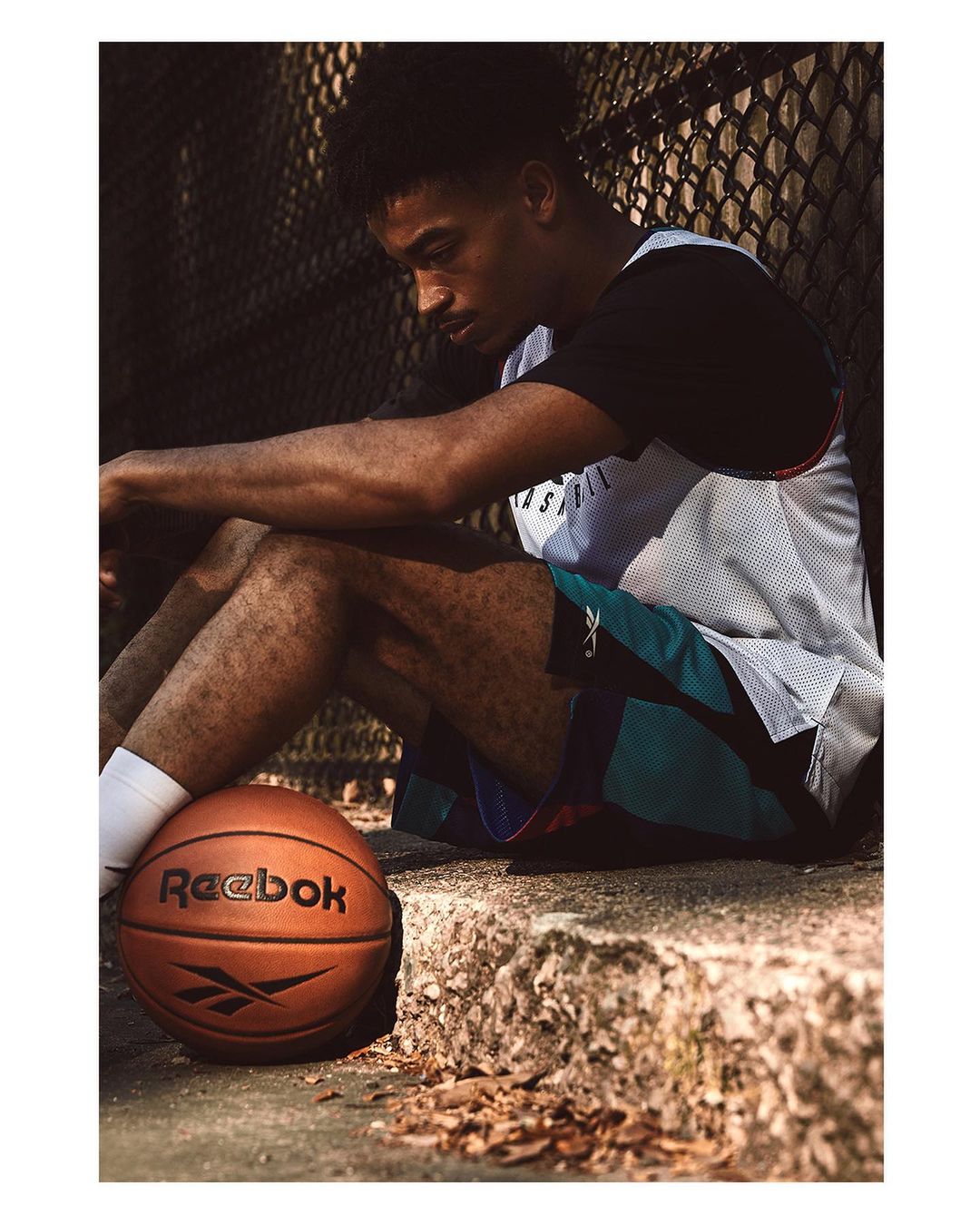 Young adult male in athletic wear seated outdoors with a basketball. He appears contemplative against a blurry urban background.