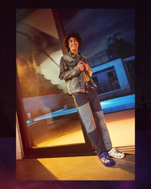 Young adult male poses in a denim jacket and patchwork jeans, wearing mismatched Crocs, beside a reflective glass wall and blue-lit building.