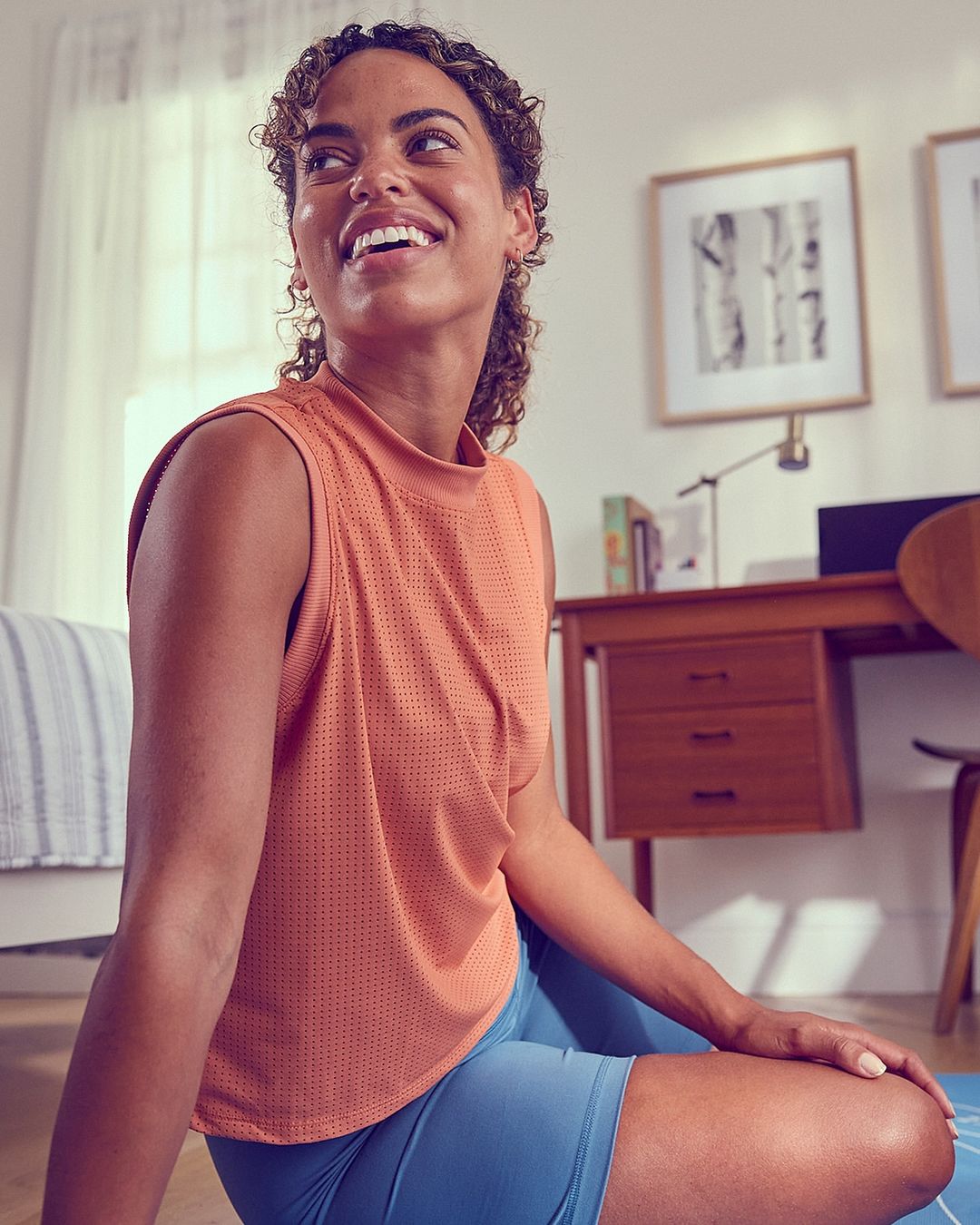 Smiling woman in fitness apparel seated on a yoga mat indoors, with a desk and framed art in the background, side lit.