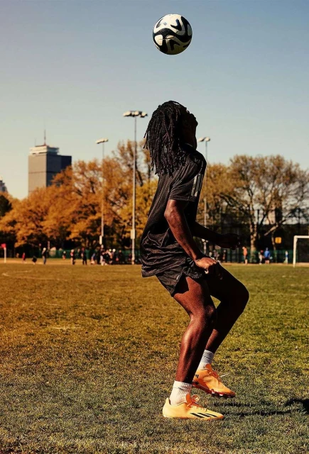 African American soccer player balances a ball on head at outdoor green field with cityscape view on a sunny day under blue skies.
