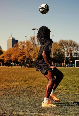 African American soccer player balances a ball on head at outdoor green field with cityscape view on a sunny day under blue skies.