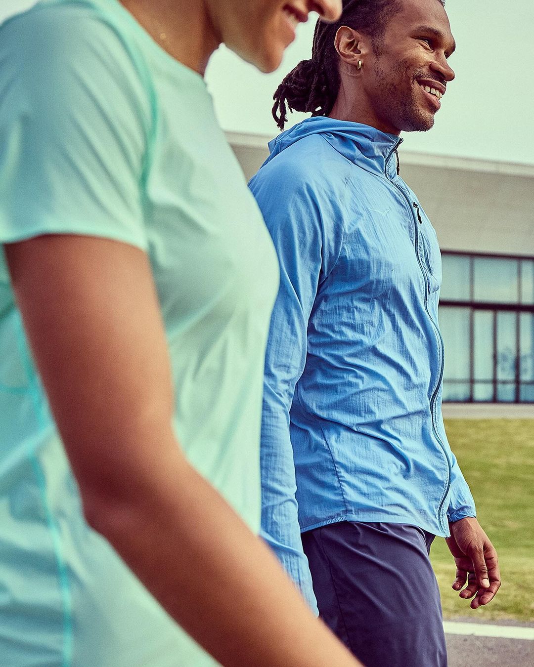 An African-American man and a woman walk side-by-side outdoors; the man is wearing a blue hooded jacket, and the woman is in a mint-green t-shirt.