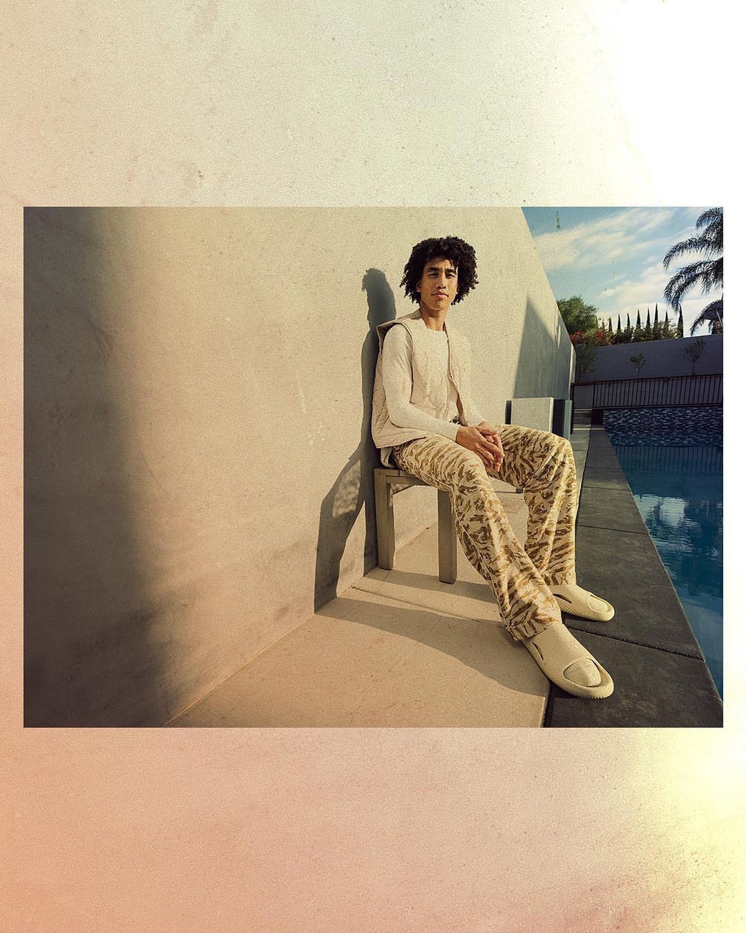 Young man with curly hair seated on a chair by a swimming pool outdoors in bright sunlight