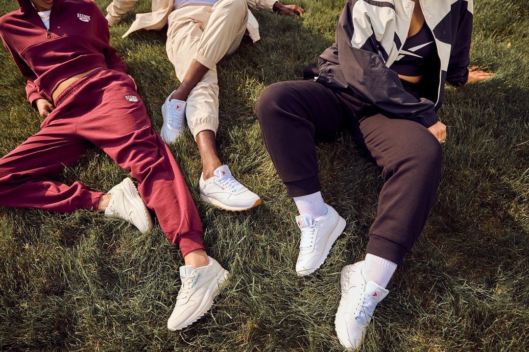 Three people are sitting on the grass wearing sneakers, sweatpants, and track jackets, in a lifestyle shot for a sports campaign.