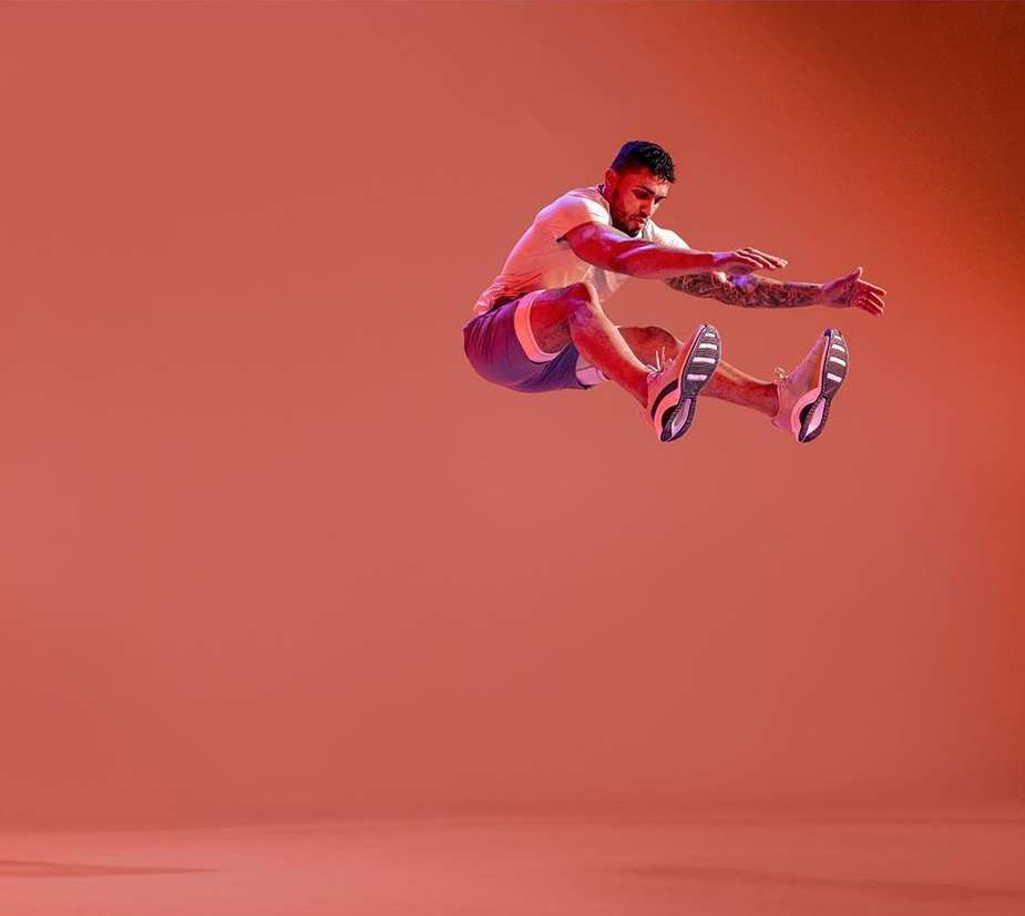 Athletic man leaps mid-air against a solid orange background, showcasing fitness and dynamic movement in a studio setting with vibrant lighting.
