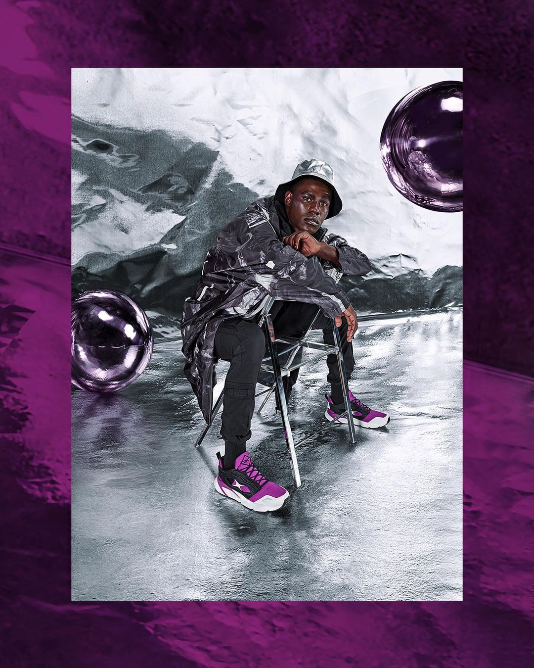 Adult male sitting on a chair in a silver and purple studio wearing a patterned jacket, matching hat, black pants and purple sneakers.