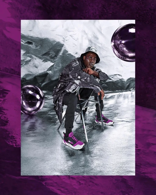 Adult male sitting on a chair in a silver and purple studio wearing a patterned jacket, matching hat, black pants and purple sneakers.