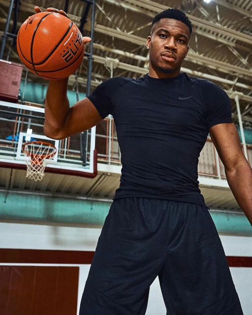 Adult male basketball player holding an orange basketball in an indoor court with a hoop and backboard.