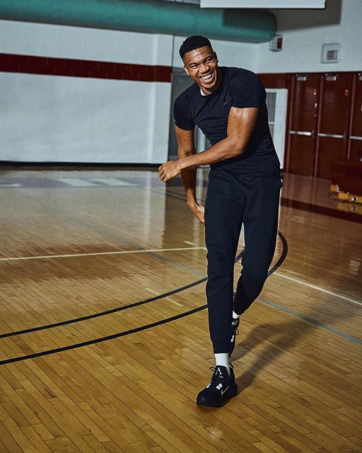 Smiling adult male athlete in sports clothing and athletic shoes stands on a basketball court with wooden flooring.
