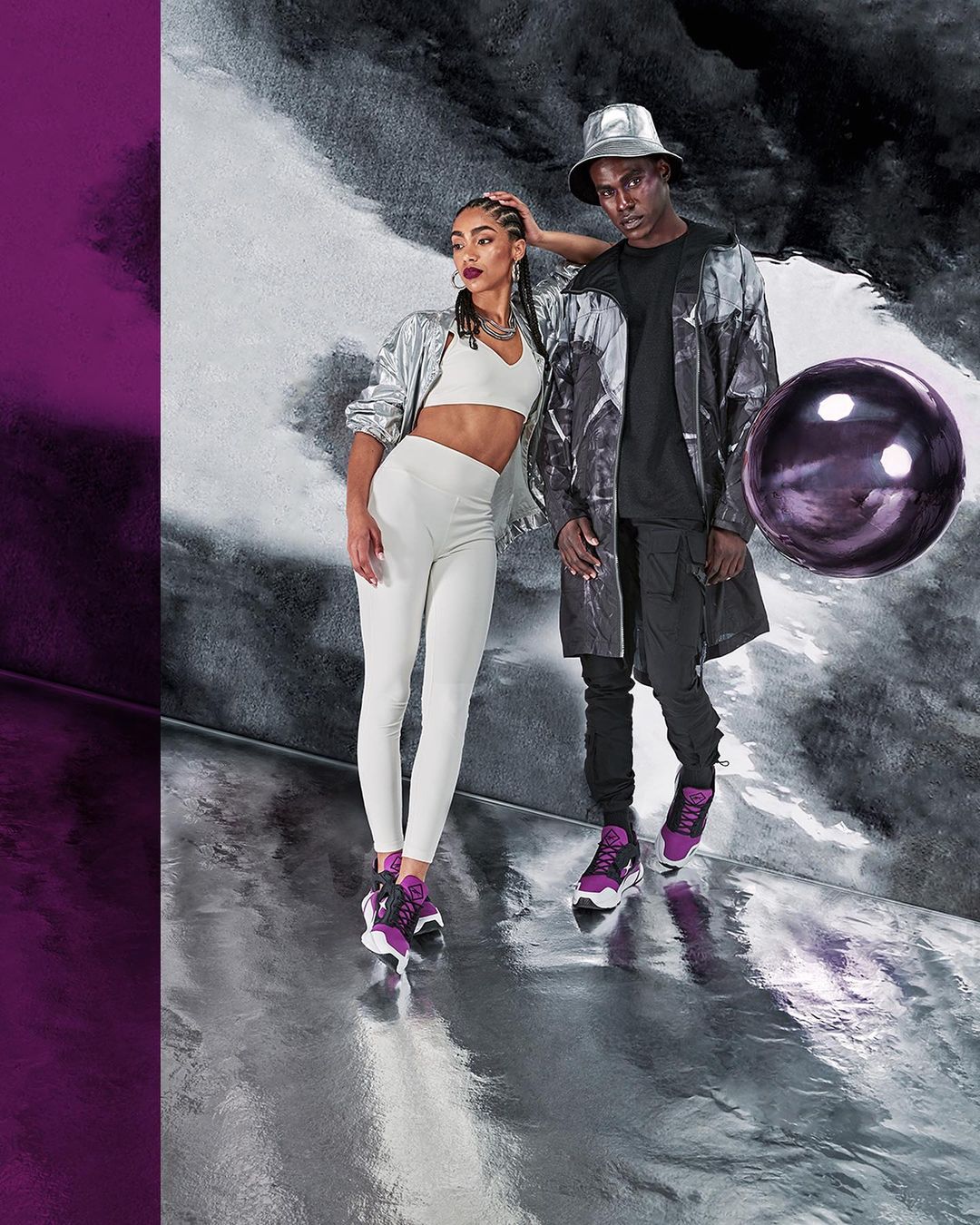 Two fashion models pose in athleticwear including sneakers in a studio setting with purple and silver reflective walls and floor.