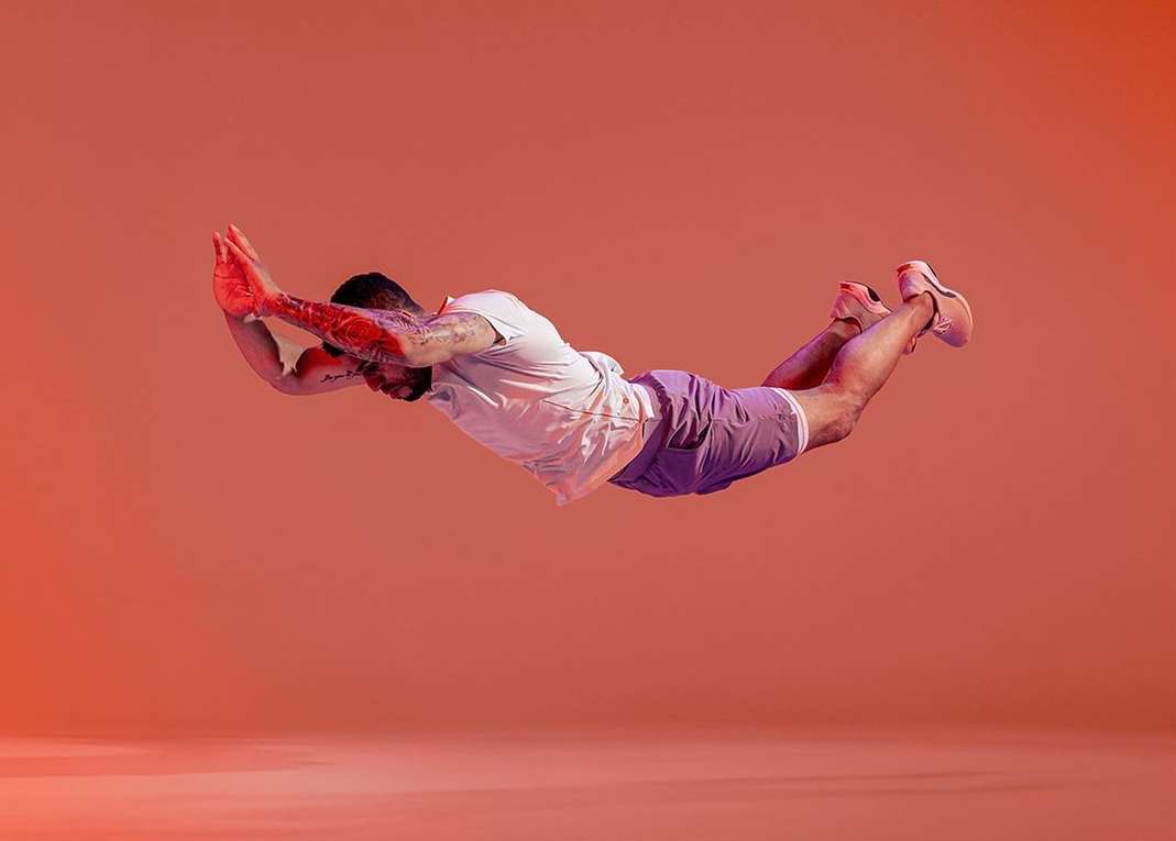 Athletic man in mid-air, arms raised, tattoos visible, wearing shorts and sneakers against a solid orange studio backdrop.