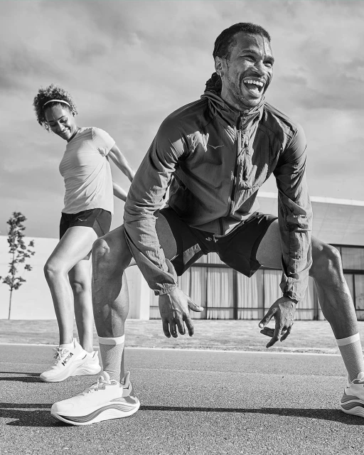 Monochrome shot of two athletes in sportswear stretching outdoors on a paved road with a modern building in the background on a sunny day.