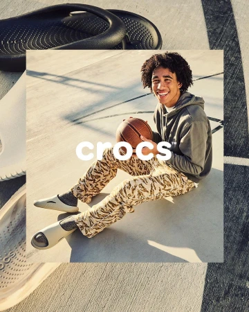 Young man smiling while sitting on an outdoor concrete court holding a basketball, wearing a grey hoodie and patterned trousers.