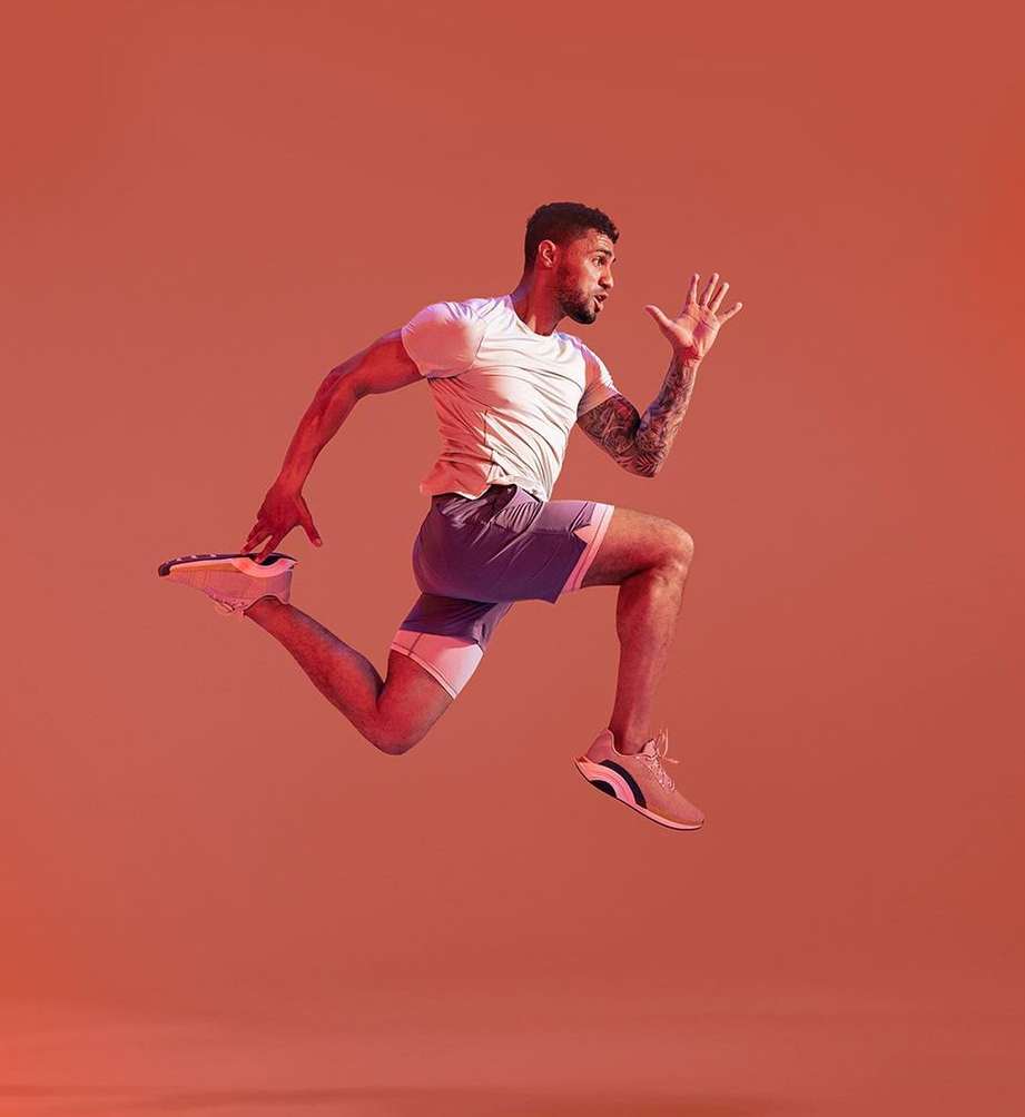 Athletic man in white shirt and gray shorts jumps mid-air with forward arm motion, captured against a vibrant orange backdrop in a fitness pose.