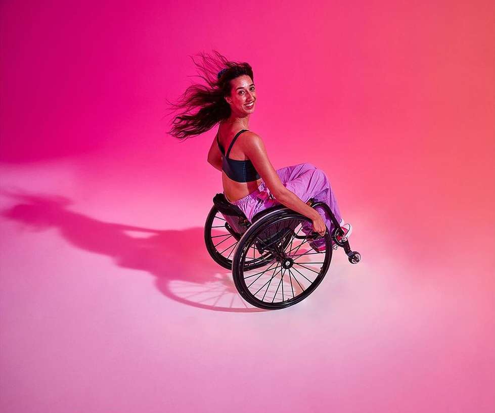 Smiling woman with long dark hair sits in a wheelchair in a photo studio with vibrant pink-to-orange gradient lighting.