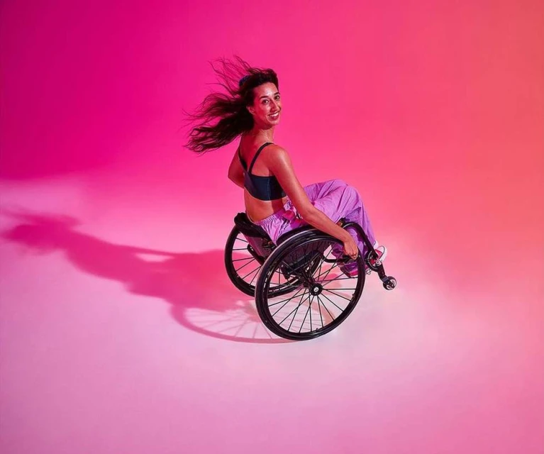 Smiling woman with long dark hair sits in a wheelchair in a photo studio with vibrant pink-to-orange gradient lighting.