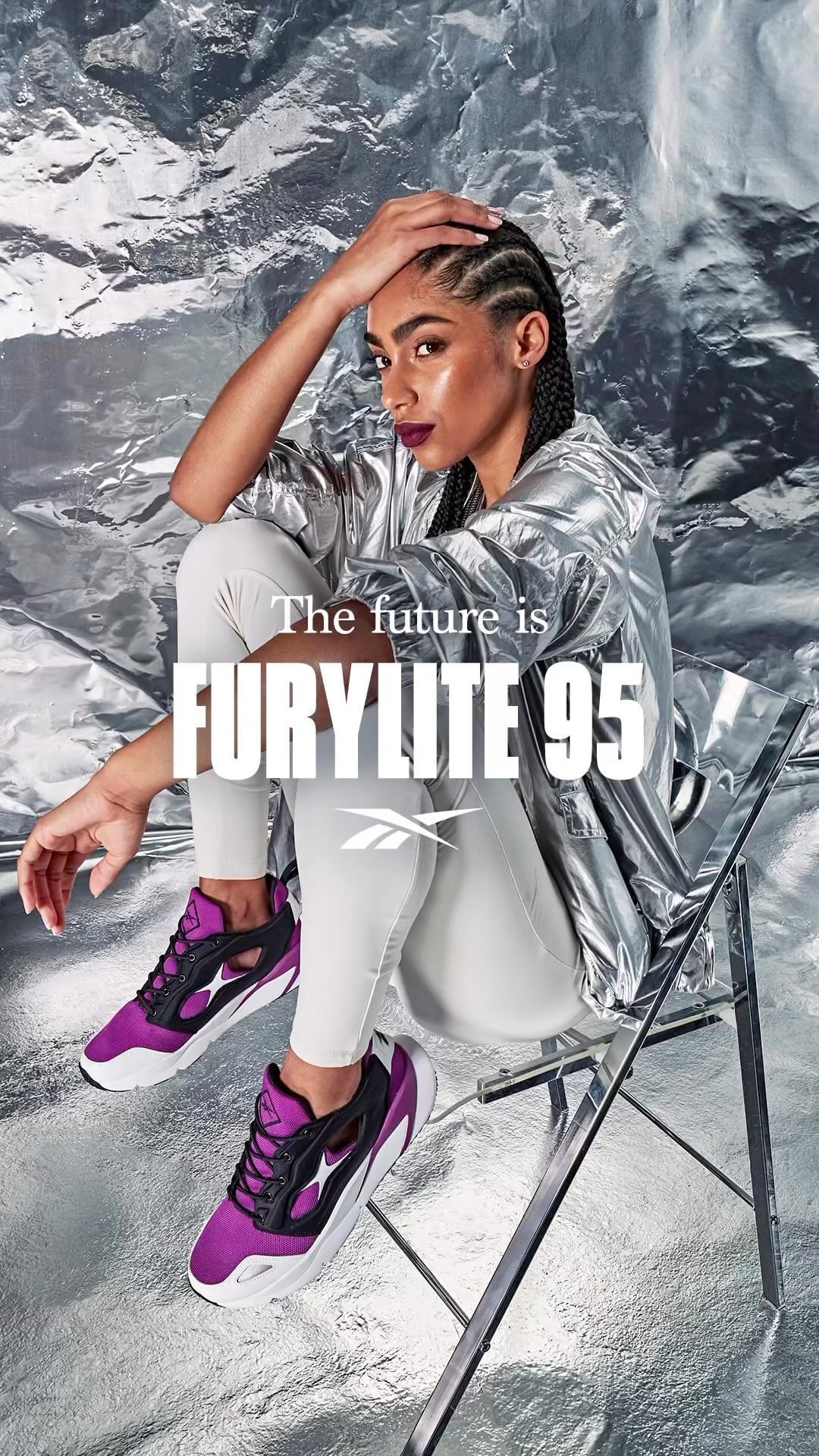 Woman in silver jacket poses with purple and black sneakers on a transparent chair against a crumpled silver background.