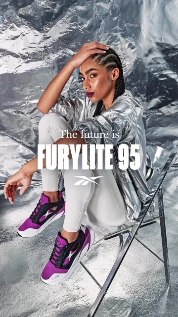 Woman in silver jacket poses with purple and black sneakers on a transparent chair against a crumpled silver background.